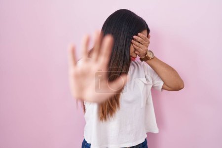 Foto de Mujer árabe joven de pie sobre fondo rosa cubriendo los ojos con las manos y haciendo gesto de parada con expresión triste y miedo. concepto avergonzado y negativo. - Imagen libre de derechos