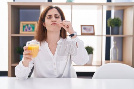 Foto de Mujer morena bebiendo vaso de jugo de naranja boca y labios cerrados como cremallera con los dedos. secreto y silencioso, tabú hablando - Imagen libre de derechos