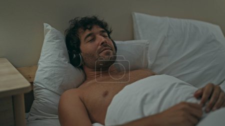 Téléchargez les photos : Jeune homme hispanique écoutant de la musique allongée sur le lit dormant dans la chambre - en image libre de droit