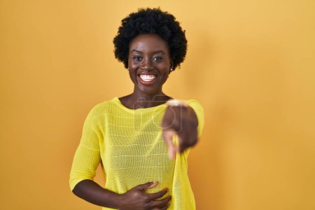 Foto de Mujer joven africana de pie sobre el estudio amarillo riéndose de ti, señalando con el dedo a la cámara con la mano sobre el cuerpo, expresión de vergüenza - Imagen libre de derechos
