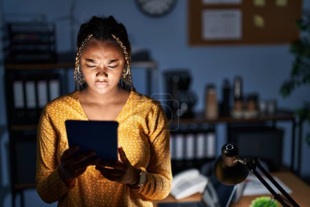 Photo for African american woman with braids working at the office at night with tablet skeptic and nervous, frowning upset because of problem. negative person. - Royalty Free Image