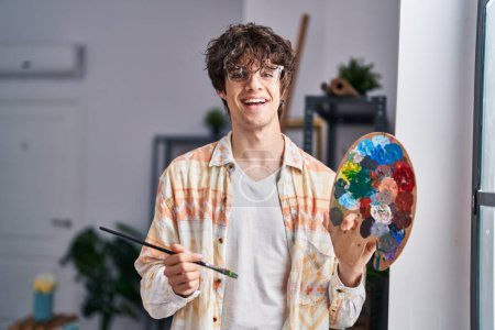 Foto de Young hispanic man artist holding paintbrush and palette at art studio - Imagen libre de derechos
