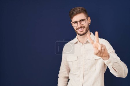 Foto de Hombre hispano con barba de pie sobre fondo azul sonriendo mirando a la cámara mostrando los dedos haciendo señal de victoria. número dos. - Imagen libre de derechos