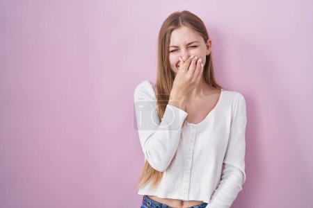 Téléchargez les photos : Jeune femme caucasienne debout sur fond rose sentant quelque chose de puant et dégoûtant, odeur intolérable, retenant souffle avec les doigts sur le nez. mauvaise odeur - en image libre de droit