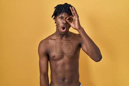 Téléchargez les photos : Jeune homme africain avec dreadlocks debout torse nu faisant geste ok choqué par le visage surpris, oeil regardant à travers les doigts. expression incrédule. - en image libre de droit