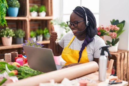 Téléchargez les photos : Jeune femme africaine travaillant au magasin de fleuriste faire appel vidéo souriant heureux et positif, pouce levé faire excellent et signe d'approbation - en image libre de droit
