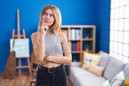 Foto de Mujer rubia caucásica de pie en la sala de estar cara seria pensando en la pregunta con la mano en la barbilla, reflexivo acerca de la idea confusa - Imagen libre de derechos