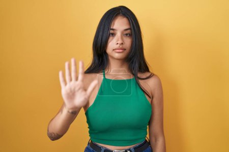 Photo for Brunette woman standing over yellow background doing stop sing with palm of the hand. warning expression with negative and serious gesture on the face. - Royalty Free Image