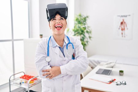Téléchargez les photos : Jeune femme médecin hispanique portant uniforme de médecin et lunettes de réalité virtuelle souriant et riant fort à haute voix parce que drôle blague folle. - en image libre de droit