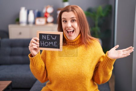 Photo for Young hispanic woman with red hair holding blackboard with new home text celebrating achievement with happy smile and winner expression with raised hand - Royalty Free Image