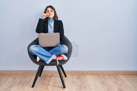 Téléchargez les photos : Jeune femme hispanique assise sur une chaise en utilisant ordinateur portable bouche et lèvres fermées comme zip avec les doigts. secret et silencieux, tabou parlant - en image libre de droit
