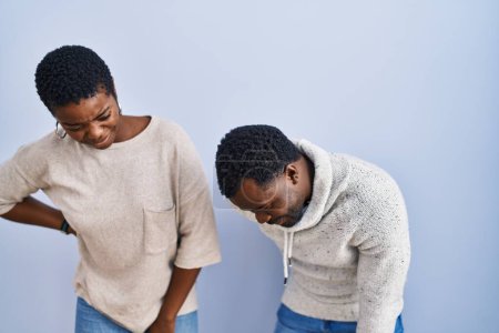 Foto de Joven pareja afroamericana de pie sobre fondo azul juntos sufriendo de dolor de espalda, tocando la espalda con la mano, dolor muscular - Imagen libre de derechos