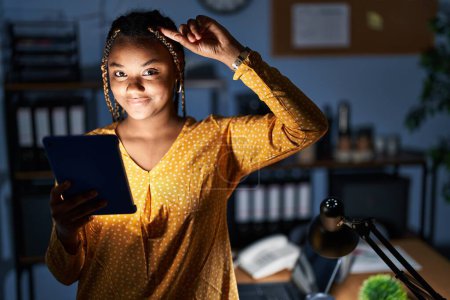 Foto de Mujer afroamericana con trenzas trabajando en la oficina por la noche con la tableta sonriendo apuntando a la cabeza con un dedo, gran idea o pensamiento, buena memoria - Imagen libre de derechos