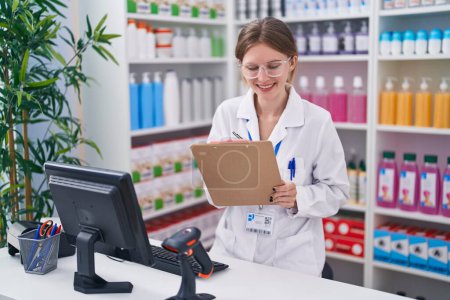 Téléchargez les photos : Jeune femme blonde pharmacienne utilisant l'écriture informatique sur le document à la pharmacie - en image libre de droit