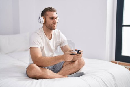Téléchargez les photos : Jeune homme caucasien assis sur le lit à la maison jouant à des jeux vidéo détendu avec une expression sérieuse sur le visage. simple et naturel regardant la caméra. - en image libre de droit