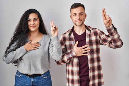 Téléchargez les photos : Jeune couple hispanique debout sur fond blanc souriant jurer avec la main sur la poitrine et les doigts vers le haut, faisant un serment de fidélité promesse - en image libre de droit