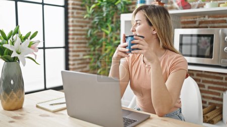 Téléchargez les photos : Jeune femme blonde utilisant un ordinateur portable buvant du café à la salle à manger - en image libre de droit