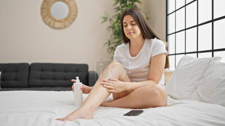 Téléchargez les photos : Jeune belle femme hispanique appliquant un traitement de la peau sur les jambes assis sur le lit dans la chambre - en image libre de droit
