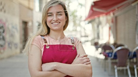 Téléchargez les photos : Jeune femme blonde serveuse souriante confiante debout avec les bras croisés geste à la terrasse du café - en image libre de droit
