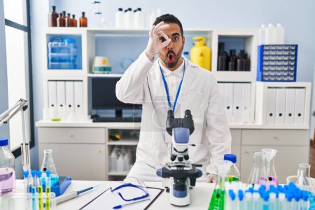 Photo for Young hispanic man with beard working at scientist laboratory doing ok gesture shocked with surprised face, eye looking through fingers. unbelieving expression. - Royalty Free Image