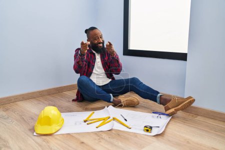 Téléchargez les photos : Afro-Américain assis sur le sol à la nouvelle maison en regardant les plans montrant du doigt la caméra avec un visage heureux et drôle. bonne énergie et vibes. - en image libre de droit