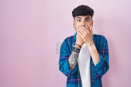 Foto de Un joven hispano con barba de pie sobre fondo rosa sorprendió cubriendo la boca con las manos por error. concepto secreto. - Imagen libre de derechos