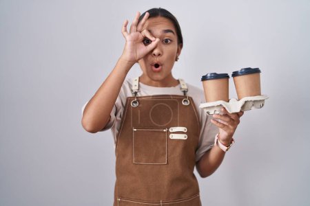 Foto de Mujer hispana joven vistiendo delantal de camarera profesional sosteniendo café haciendo un gesto bien sorprendido con la cara sorprendida, los ojos mirando a través de los dedos. expresión incrédula. - Imagen libre de derechos