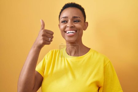 Téléchargez les photos : Afro-américaine femme souriant confiant faire ok signe avec pouce vers le haut sur fond jaune isolé - en image libre de droit