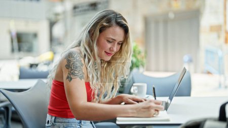 Photo for Young blonde woman using laptop taking notes smiling at coffee shop terrace - Royalty Free Image