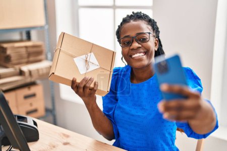 Téléchargez les photos : African american woman ecommerce business worker make selfie by smartphone holding package at office - en image libre de droit