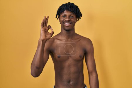 Foto de Joven hombre africano con rastas de pie sin camisa sonriendo positiva haciendo signo ok con la mano y los dedos. expresión exitosa. - Imagen libre de derechos
