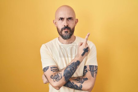 Téléchargez les photos : Homme hispanique avec des tatouages debout sur fond jaune pointant avec le doigt de la main sur le côté montrant la publicité, visage sérieux et calme - en image libre de droit