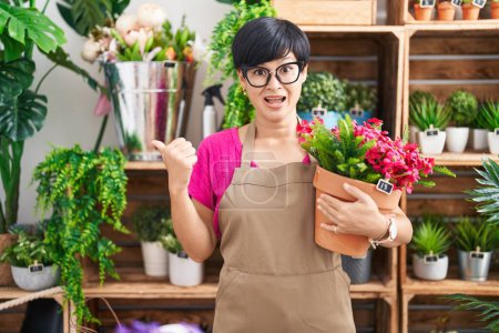 Foto de Joven mujer asiática con pelo corto trabajando en floristería sosteniendo planta apuntando pulgar hacia el lado sonriendo feliz con la boca abierta - Imagen libre de derechos