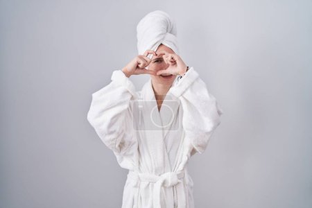 Photo for Blonde caucasian woman wearing bathrobe doing heart shape with hand and fingers smiling looking through sign - Royalty Free Image