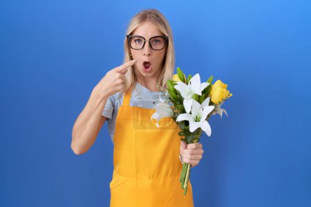Foto de Mujer joven caucásica vistiendo delantal florista sosteniendo flores sorprendidas señalando con el dedo hacia un lado, boca abierta expresión asombrada. - Imagen libre de derechos