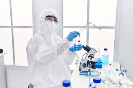 Foto de Joven científica rubia vistiendo uniforme de seguridad vertiendo líquido en el tubo de ensayo en el laboratorio - Imagen libre de derechos