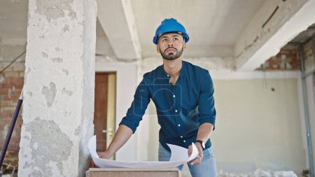 Photo for Young hispanic man architect wearing hardhat looking at blueprints at construction site - Royalty Free Image