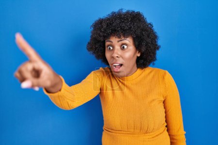Téléchargez les photos : Femme noire avec les cheveux bouclés debout sur fond bleu pointant avec le doigt surpris devant, bouche ouverte expression étonnante, quelque chose sur le devant - en image libre de droit