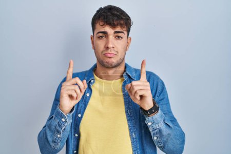 Téléchargez les photos : Jeune homme hispanique debout sur fond bleu pointant vers le haut regardant triste et contrarié, indiquant la direction avec les doigts, malheureux et déprimé. - en image libre de droit