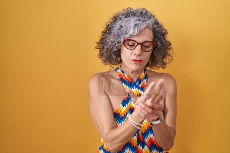 Téléchargez les photos : Femme d'âge moyen avec les cheveux gris debout sur fond jaune souffrant de douleur aux mains et aux doigts, inflammation de l'arthrite - en image libre de droit