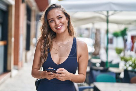 Téléchargez les photos : Jeune belle femme hispanique souriant confiant en utilisant smartphone à la terrasse du café - en image libre de droit