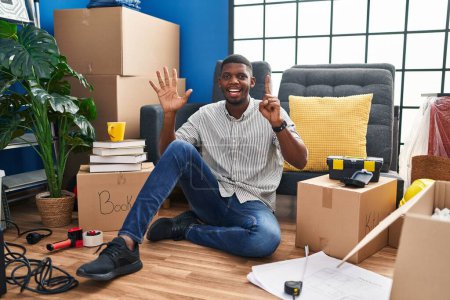 Téléchargez les photos : Afro-Américain assis sur le sol à la nouvelle maison montrant et pointant vers le haut avec les doigts numéro six tout en souriant confiant et heureux. - en image libre de droit