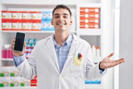 Téléchargez les photos : Bel homme hispanique travaillant à la pharmacie pharmacie montrant écran smartphone célébrant la réalisation avec sourire heureux et expression gagnante avec la main levée - en image libre de droit