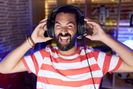 Foto de Hombre hispano con barba jugando videojuegos con auriculares sonriendo y riendo en voz alta porque divertido chiste loco. - Imagen libre de derechos