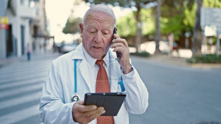 Photo for Senior grey-haired man doctor using touchpad talking on smartphone at street - Royalty Free Image