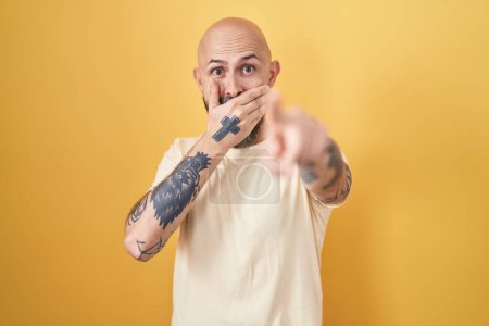 Foto de Hombre hispano con tatuajes de pie sobre fondo amarillo riéndose de ti, señalando con el dedo a la cámara con la mano sobre la boca, expresión de vergüenza - Imagen libre de derechos