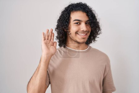 Foto de Hombre hispano con el pelo rizado de pie sobre fondo blanco renunciando a decir hola feliz y sonriente, gesto de bienvenida amistoso - Imagen libre de derechos