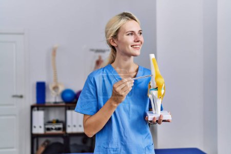 Téléchargez les photos : Jeune femme blonde pysiothérapeute souriant confiant pointant vers le modèle anatomique du genou à la clinique de réadaptation - en image libre de droit
