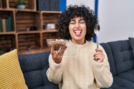 Téléchargez les photos : Jeune femme brune aux cheveux bouclés mangeant des céréales saines à grains entiers avec cuillère collant la langue heureuse avec une expression drôle. - en image libre de droit