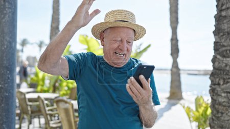 Téléchargez les photos : Homme âgé aux cheveux gris touriste portant un chapeau d'été en utilisant un smartphone avec expression gagnante au bord de la mer - en image libre de droit